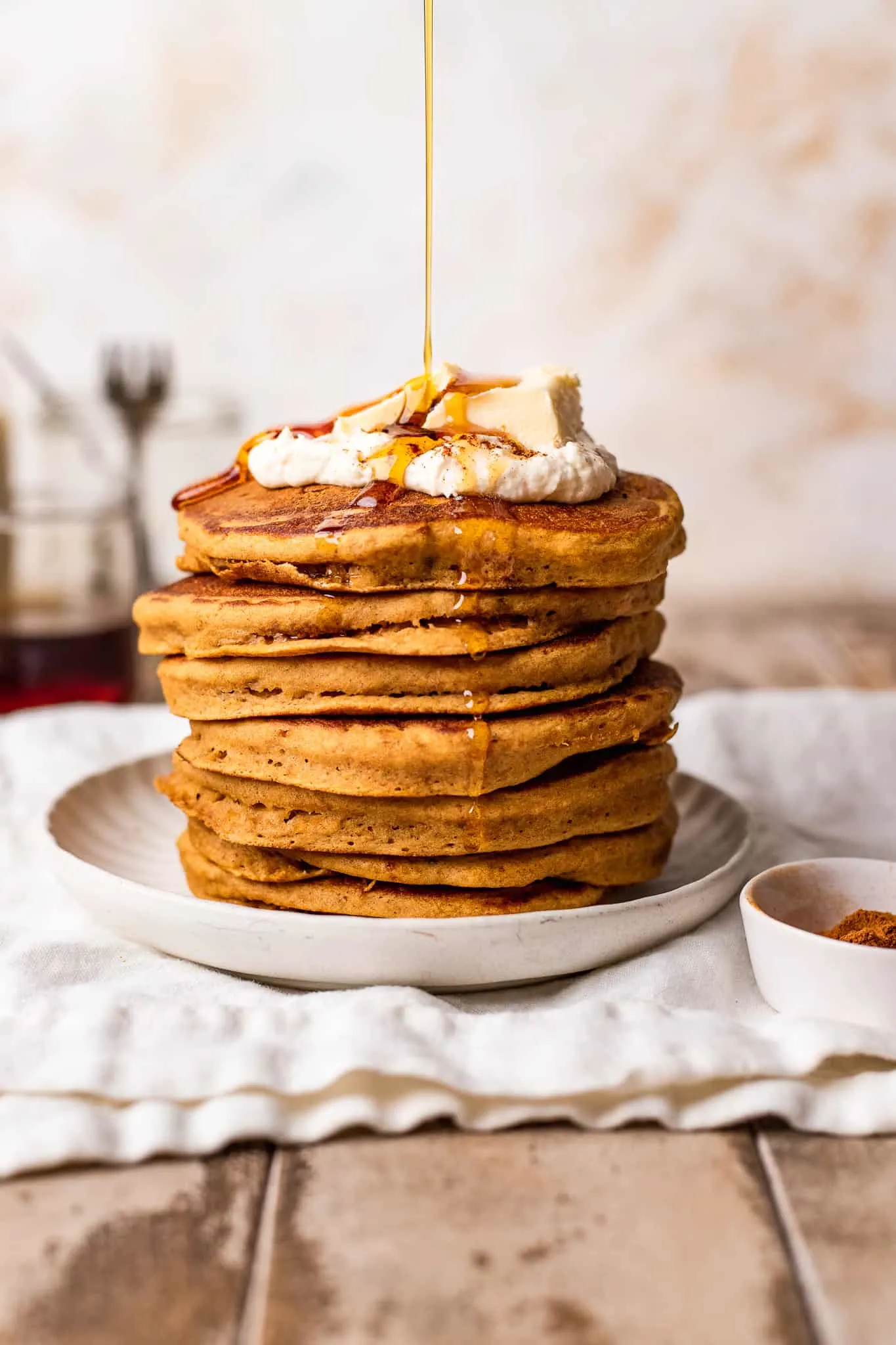 A syrup being drizzled on a stack of vegan pumpkin pancake with whipped cream and butter.