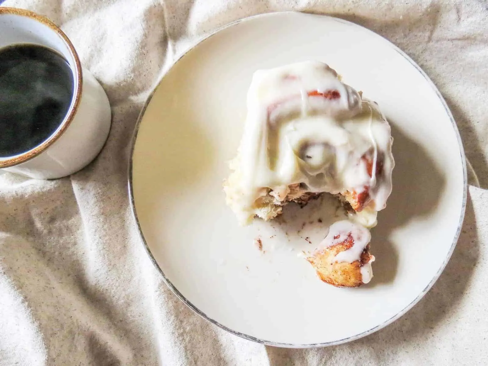 Overnight cinnamon rolls with cream cheese icing next to a cup of hot coffee.
