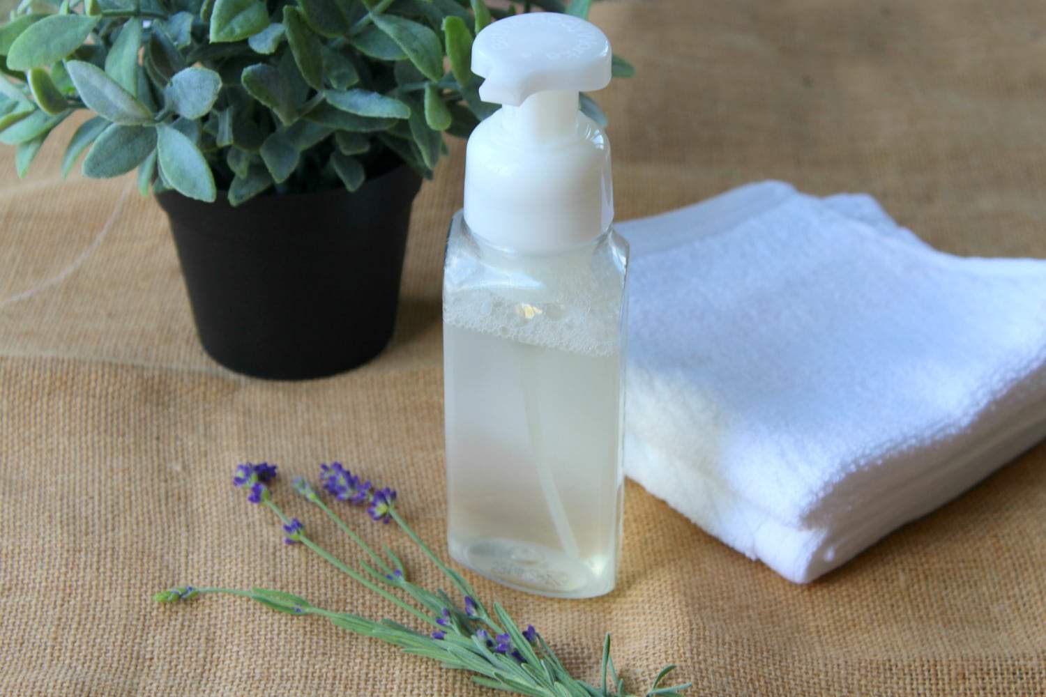 Homemade foaming hand soap next to a pot of plant, white towels and a stem of plant with purple flowers.