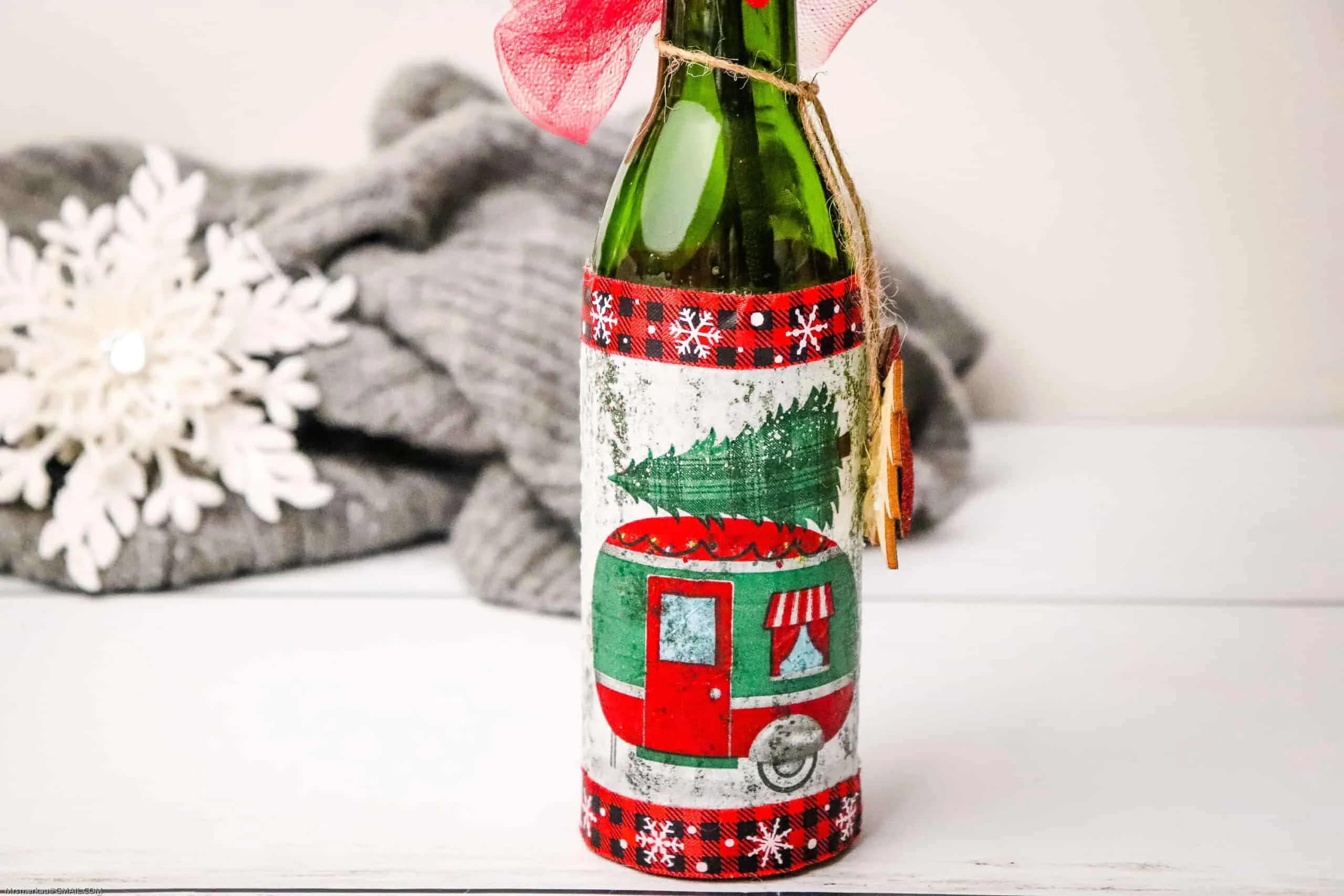 A wine bottle decorated with a Christmas tree on top of a camper. A snowflake decor and gray blanket on the background.