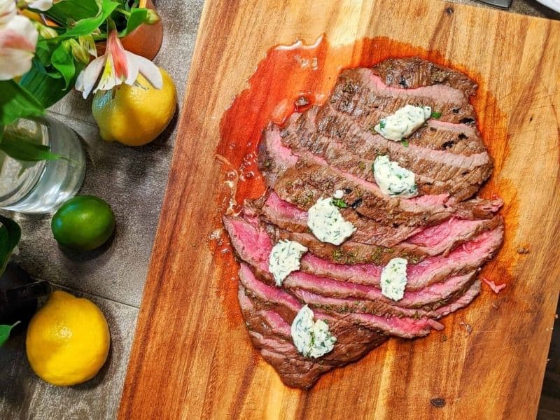 Sliced steak with herb butter on a wooden chopping board next to vases, lemon and lime.