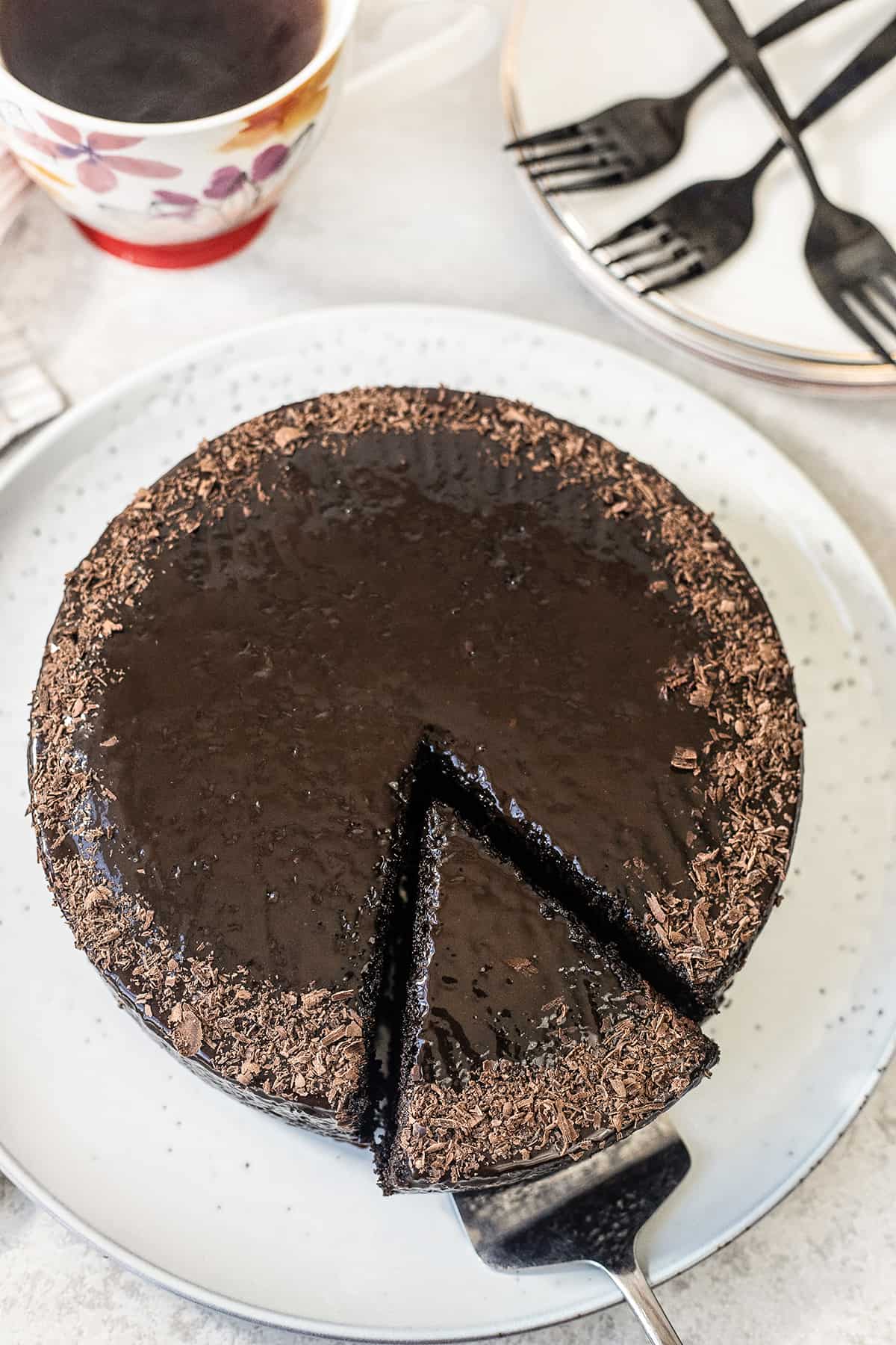 A whole chocolate cake with a slice that is about to be served next to plates with fork and a cup of coffee.