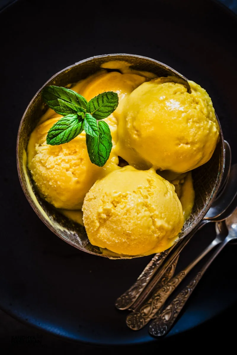 Scoops of mango ice cream in a bowl next to spoons with dark background.