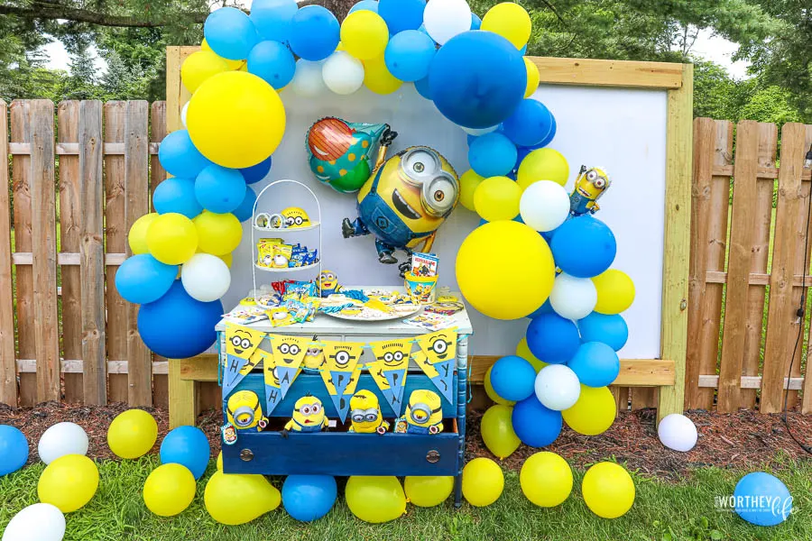Minion party table with a minion ballon and party banners. Blue and yellow balloon arc as background with same colored balloons scattered on the ground.