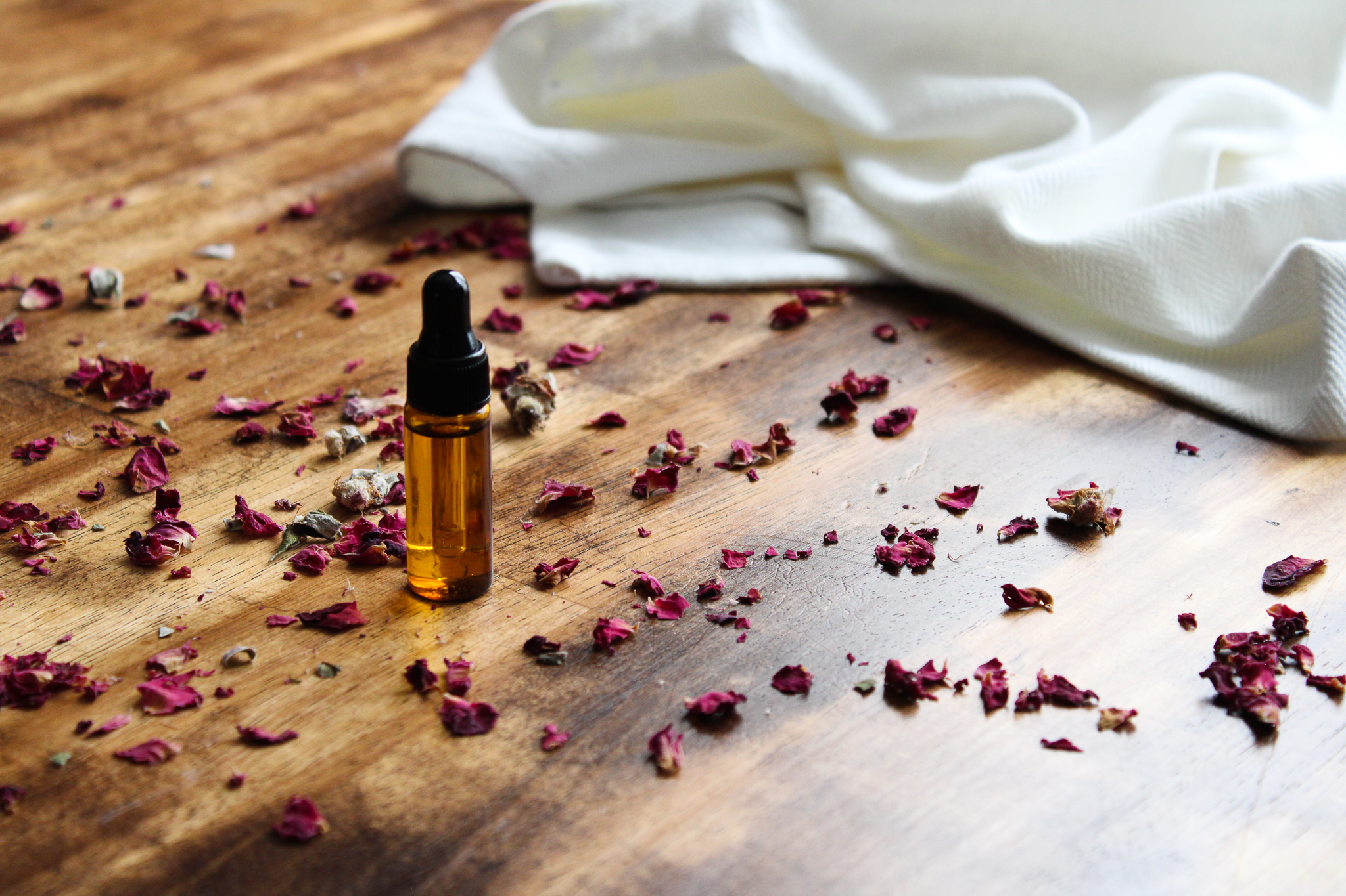 Cuticle oil dropper bottle on the wooden floor surrounded by dried rose petals next to a white cloth.