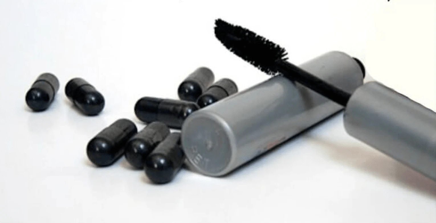A mascara brush applicator removed from the tube next to charcoal capsules.