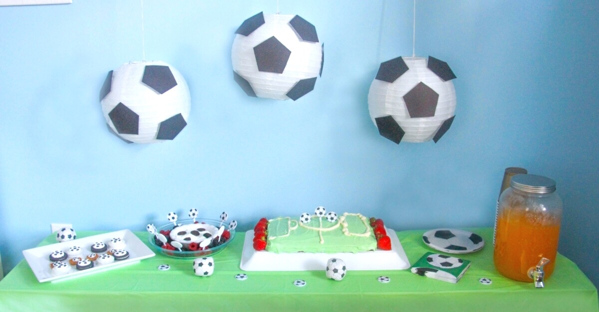 Soccer birthday party snack table with cake, cupcakes and an orange juice. Paper soccer ball decoration hanging from the top.
