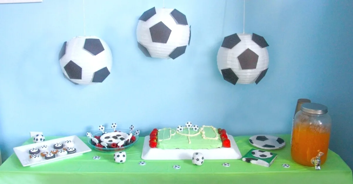 Soccer birthday party snack table with cake, cupcakes and an orange juice. Paper soccer ball decoration hanging from the top.