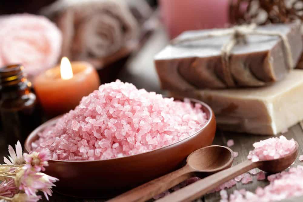 Himalayan salt in a wooden bowl next to two wooden spoons with two bar of soaps stacked together and a small lit pink candle at the background.