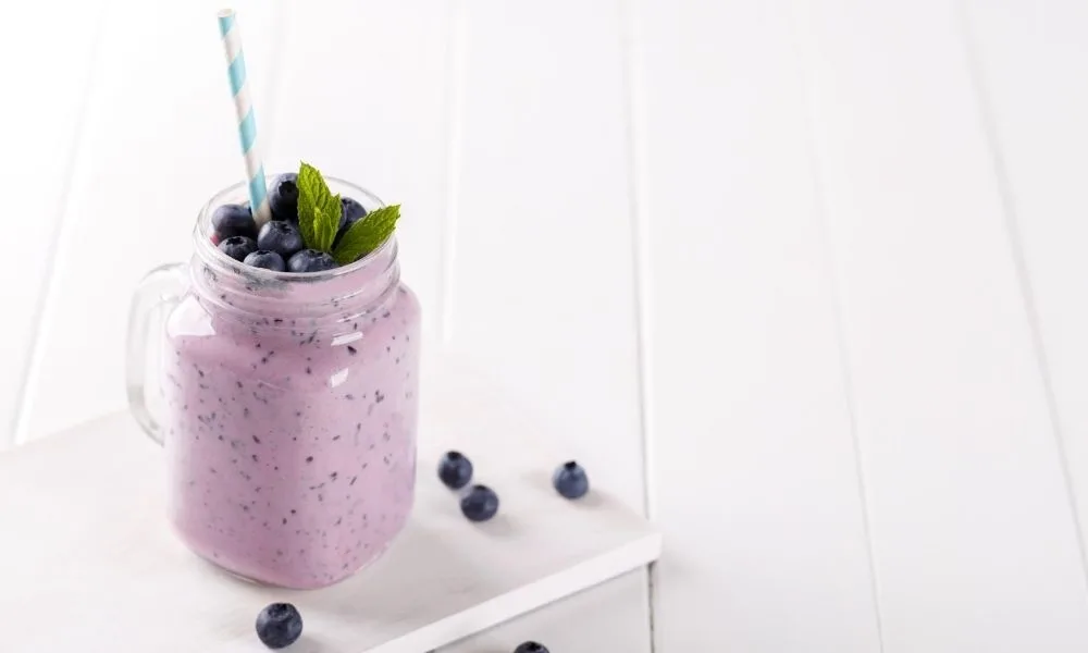 Blueberry smoothie in a mason jar with fresh blueberries and mint garnish and a white and blue stripe paper straw.