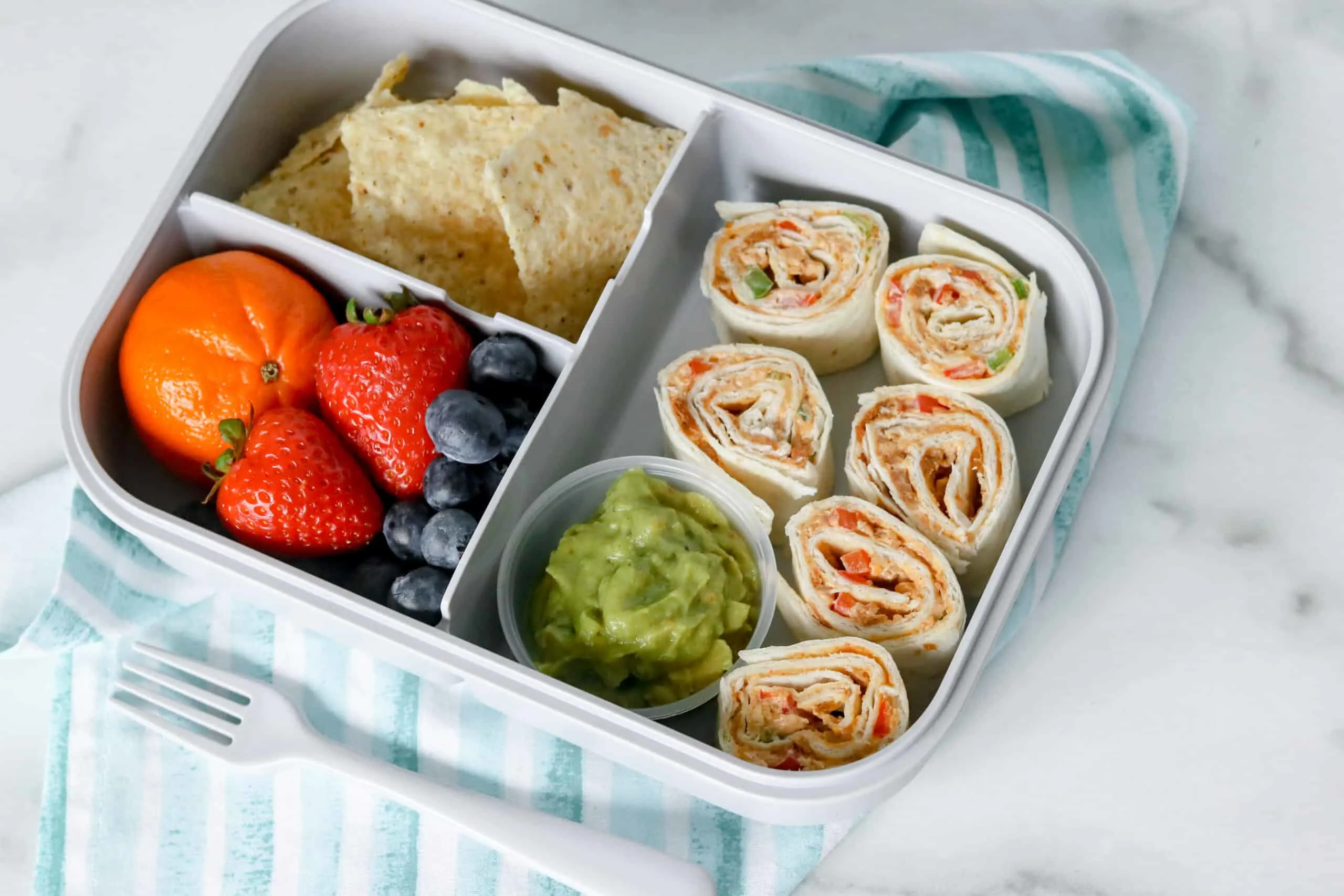 A bento box full with taco pinwheels, blueberries, strawberries, a small orange and some chips.
