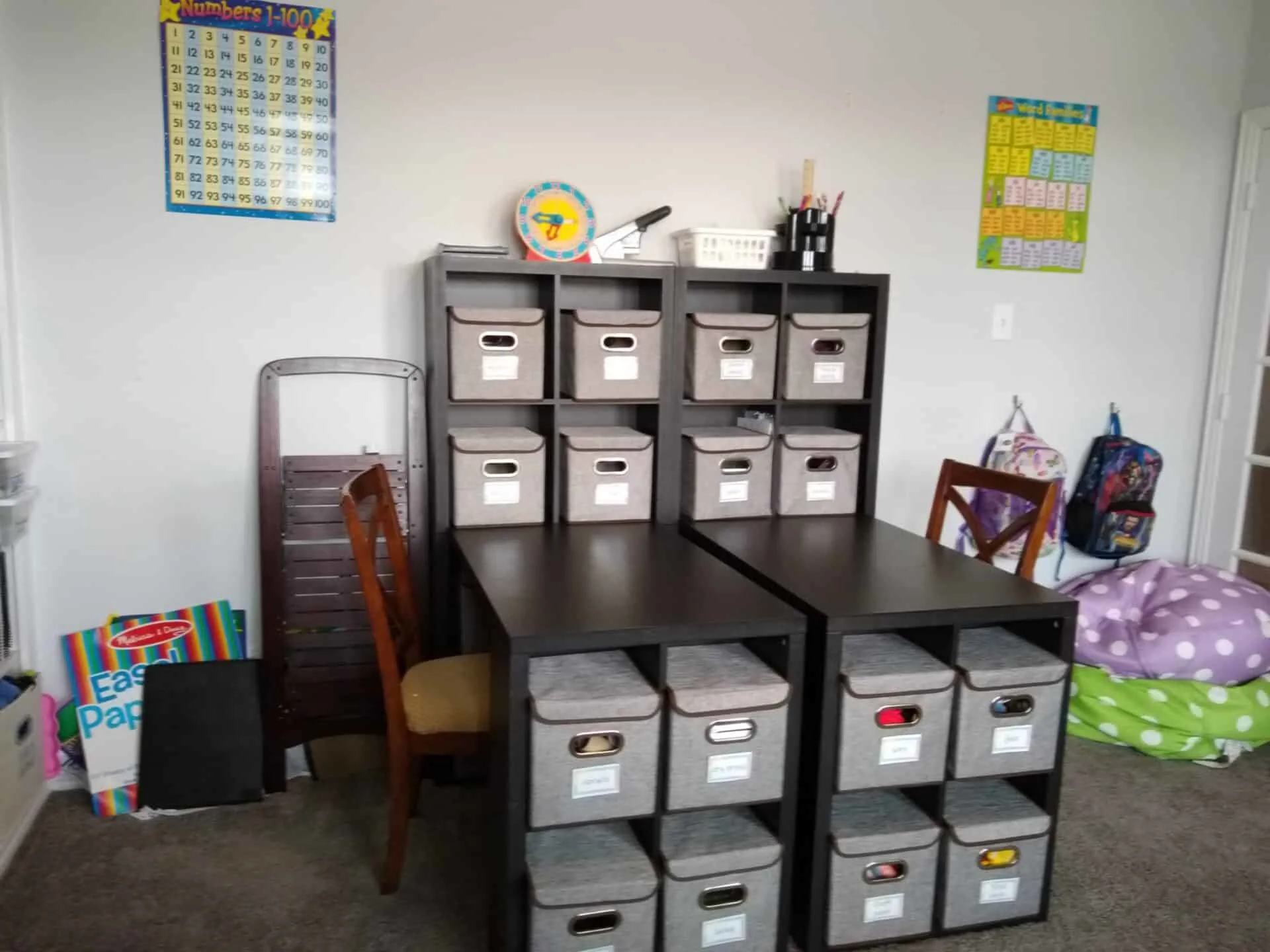 Homeschool organization shelves with grey crates that have labels on them. Two wooden study tables next to each other with two wooden chairs facing each other.