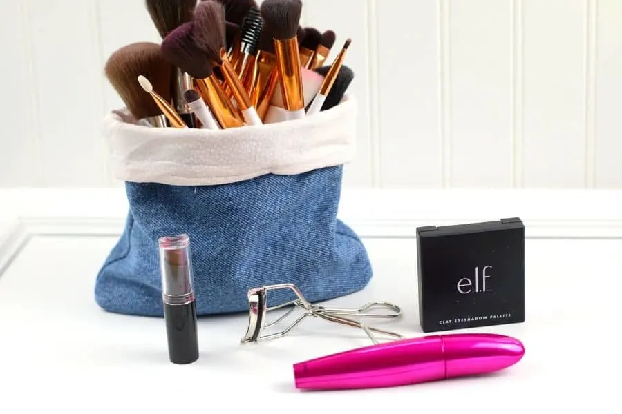 Different make up brushes coming out of a denim make up storage bag next to a lipstick, eyelash curler, mascara and face powder.