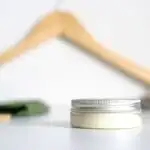 A small jar of beard balm on a white surface with a wooden hanger on the background.