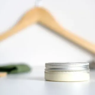 A small jar of beard balm on a white surface with a wooden hanger on the background.