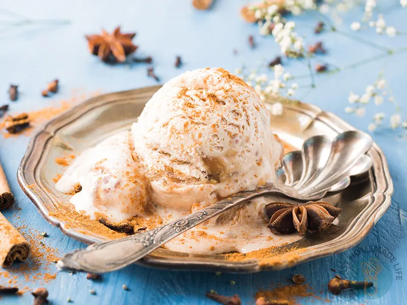 A scoop of chai ice cream garnished with cinnamon powder in a silver plate surrounded by cinnamon sticks.
