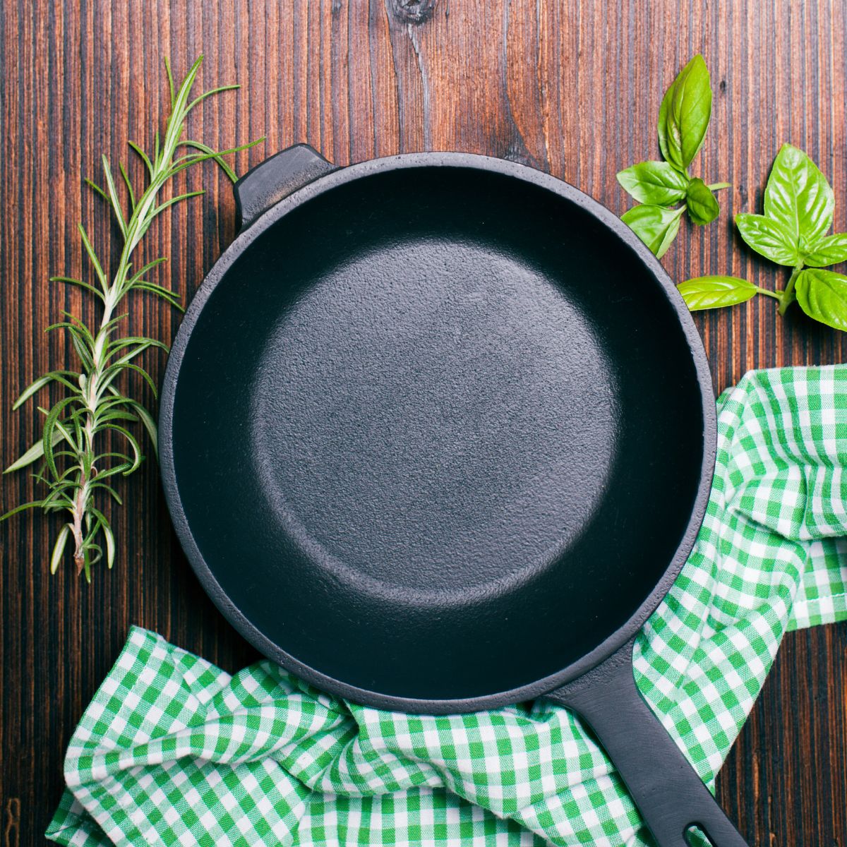 A clean cast iron skillet with green checkered kitchen towel and herbs.