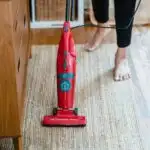 Barefoot woman on tan carpet vacuuming with red vacuum.