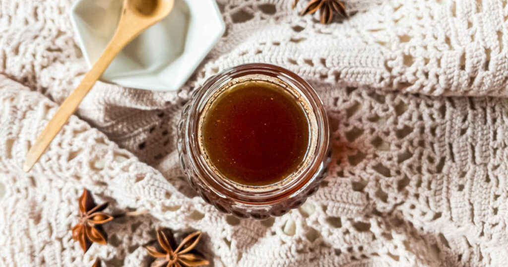 Chai syrup in a round glass bottle next to a white bowl with wooden spoon with crochet fabric background.