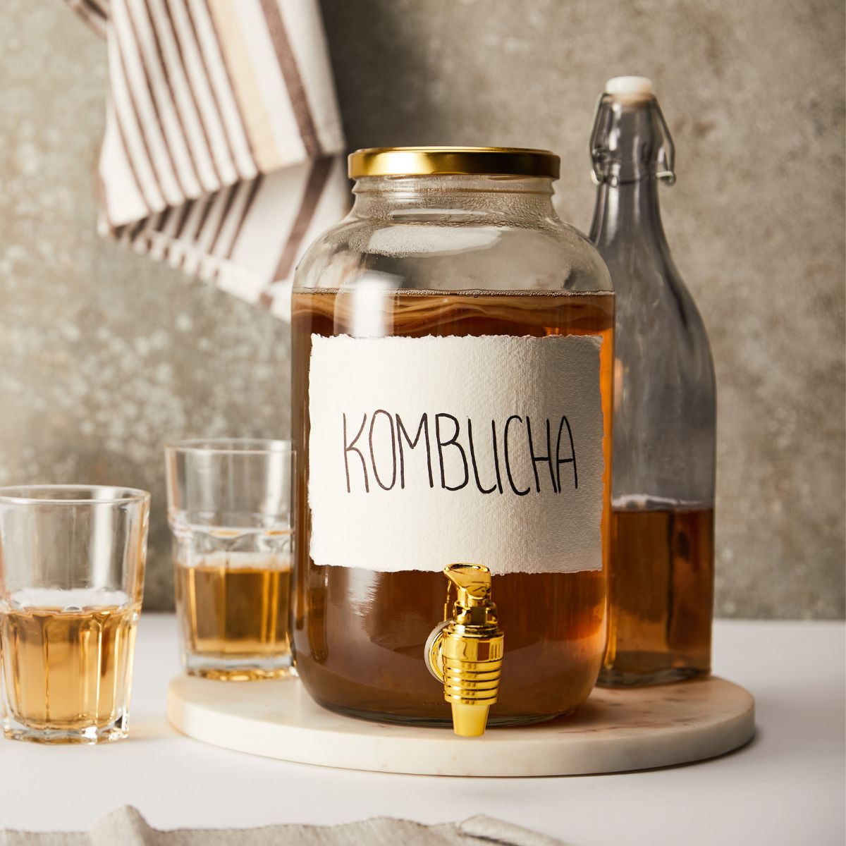 A glass dispenser of kombucha with golden faucet next to a glass bottle with brown liquid and two half filled glasses with the same liquid drink.