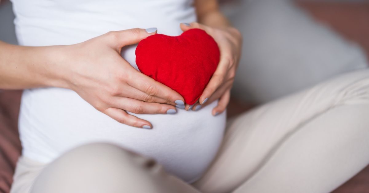 A pregnant woman holding a red heart over her growing belly.