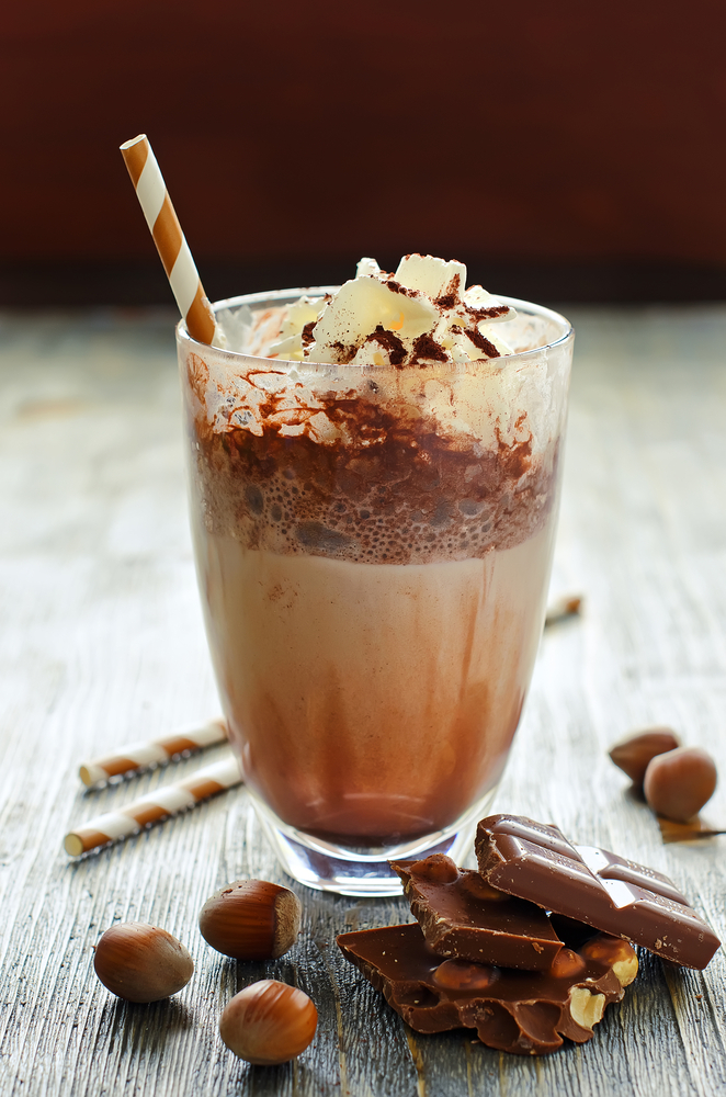 A coffee drink with a straw surrounded by chocolate and nuts on a wooden table.