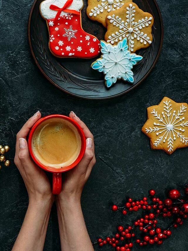 A woman holds a cup of coffee next to a plate of cookies and snowflakes.