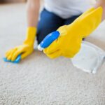 Person in yellow gloves cleaning a carpet with a spray bottle and cloth.