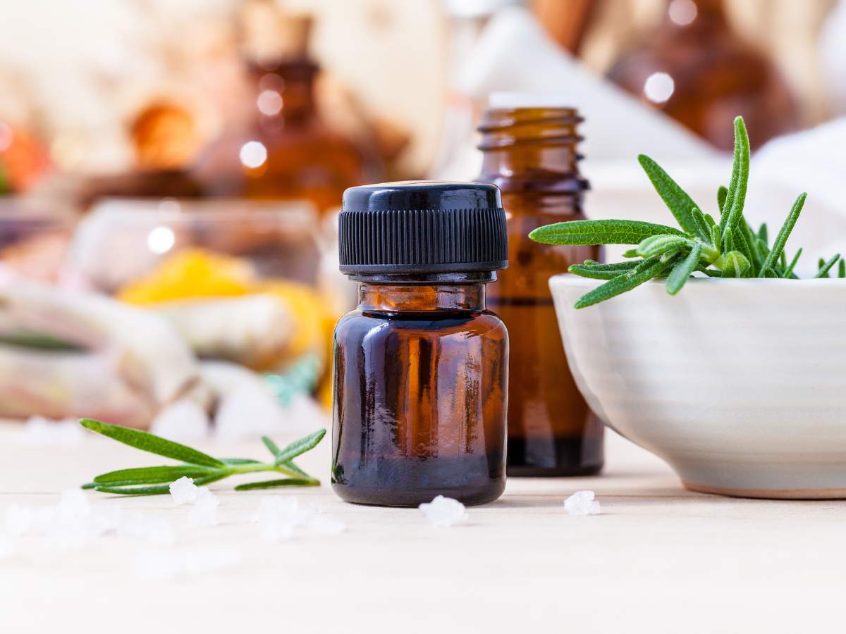 A bottle of essential oil with fresh rosemary and other herbs in the background.