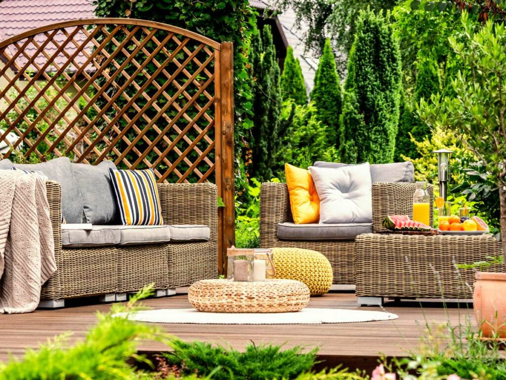 A cozy outdoor seating area featuring wicker furniture with cushions, a rug, and surrounded by lush greenery and a wooden lattice in the background.