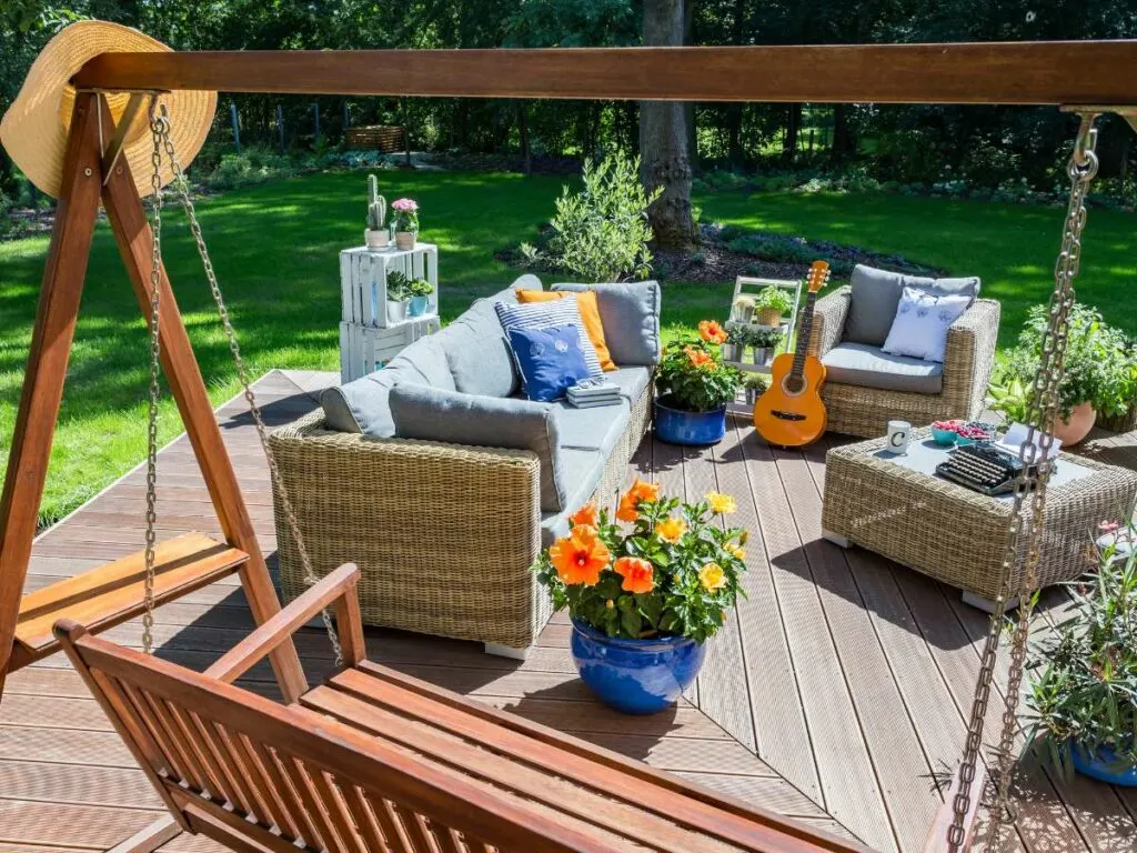 Wooden deck with a swing, wicker furniture with cushions, a guitar, and potted flowers in a sunny garden setting.