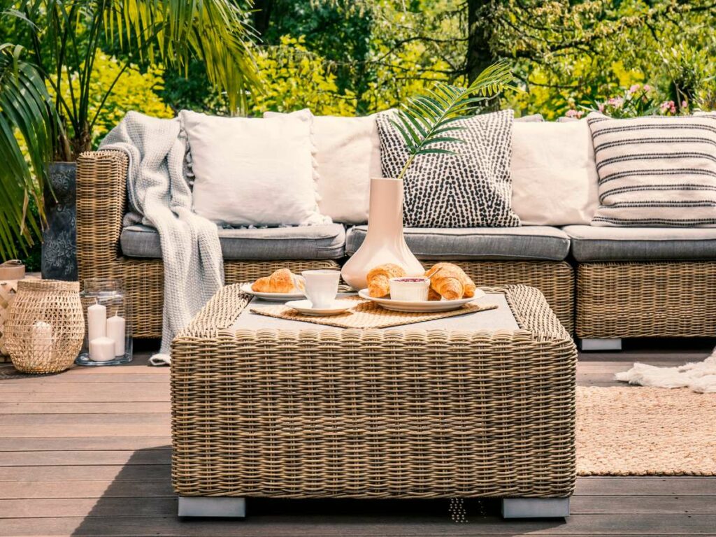 Outdoor patio setting with a wicker sofa, cushions, and a coffee table holding a vase and croissants, surrounded by lush greenery.