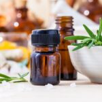 A small brown glass bottle with essential oil next to a bowl of fresh rosemary and scattered salt crystals on a wooden surface.