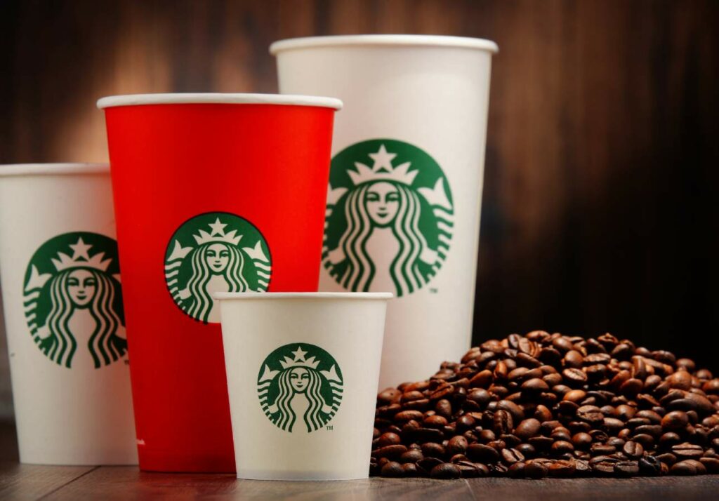 Three starbucks cups of varying sizes on a wooden surface with roasted coffee beans scattered in the foreground.