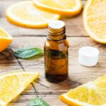 A small amber bottle of essential oil sits on a wooden table surrounded by fresh orange slices and green mint leaves.