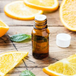 A small amber bottle of essential oil sits on a wooden table surrounded by fresh orange slices and green mint leaves.