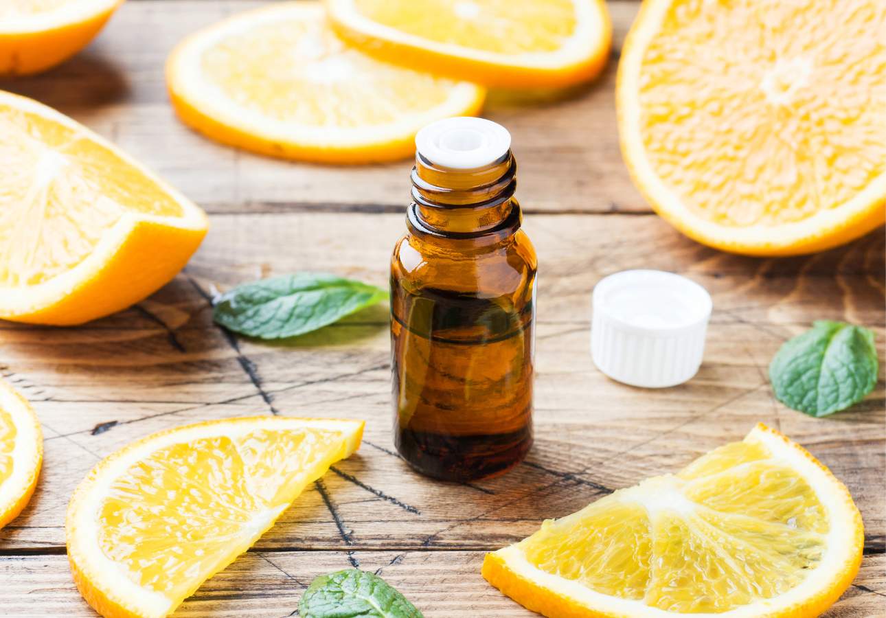 A small amber bottle of essential oil sits on a wooden table surrounded by fresh orange slices and green mint leaves.