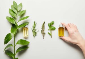 A hand holding a small bottle of yellow oil, next to three sprigs of herbs and another small bottle of yellow oil, all arranged on a white surface.