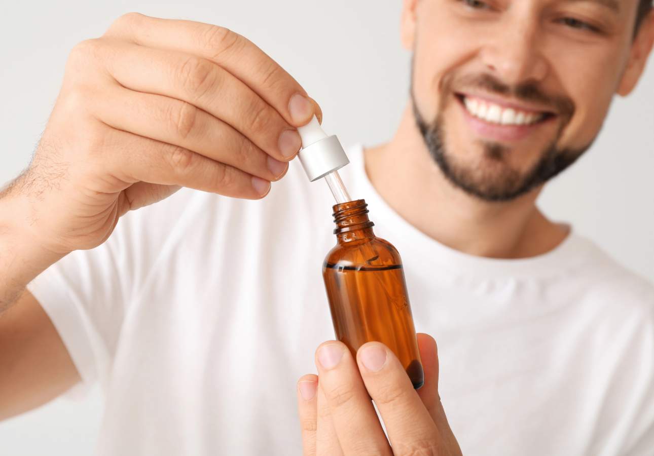 A man in a white shirt holds a brown glass dropper bottle, smiling as he uses the dropper.