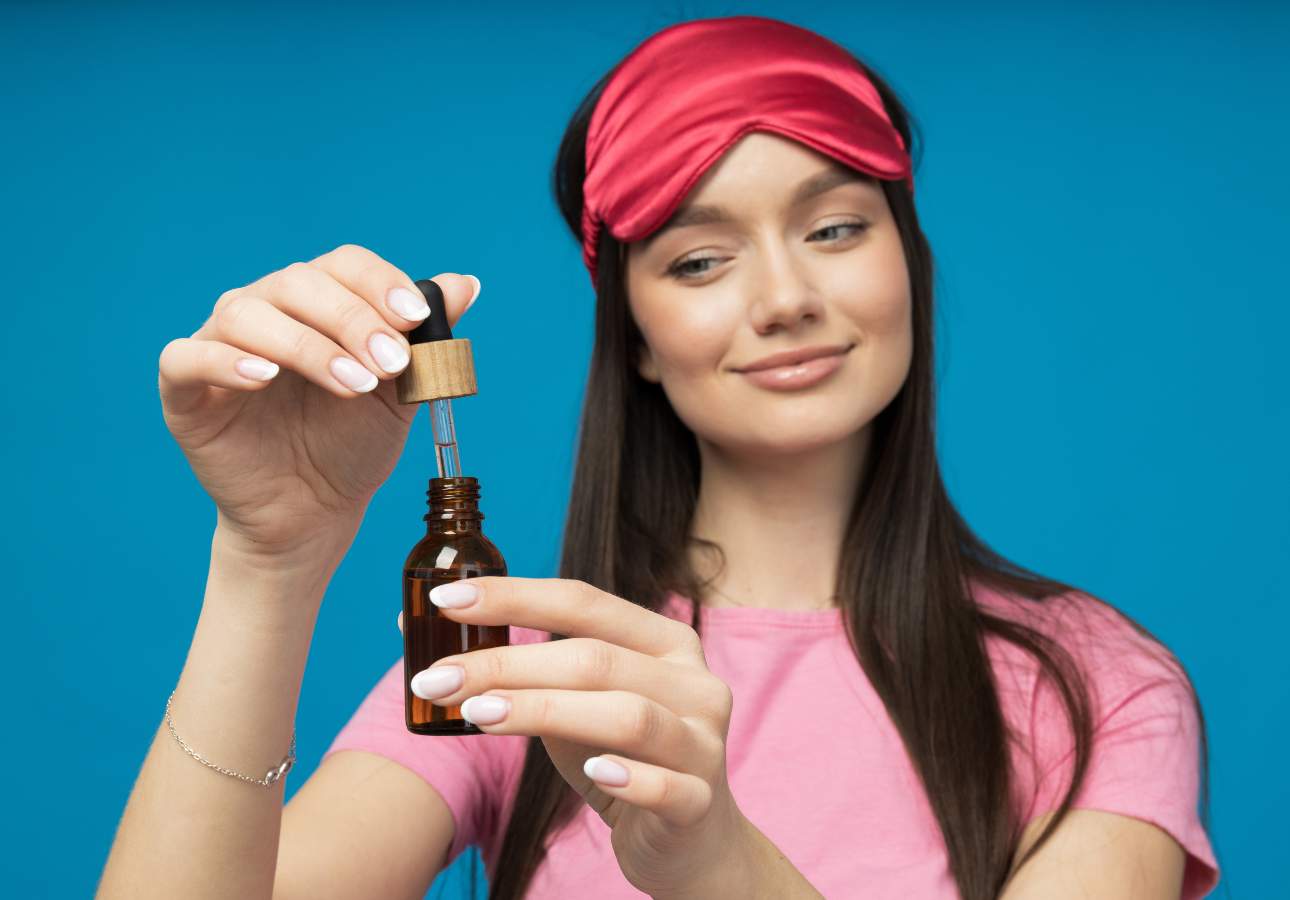 A woman in a pink sleep mask and pink shirt holds and uses a dropper bottle filled with liquid, set against a blue background.