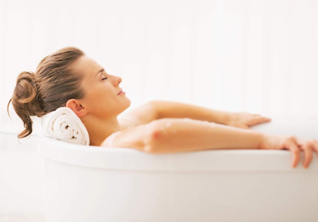 A woman leans back in a bathtub with a towel under her head, eyes closed, and appears relaxed.