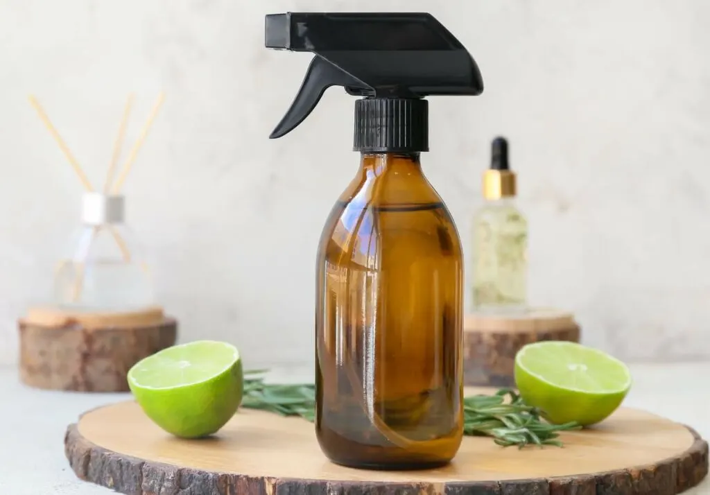 A brown spray bottle sits on a wooden board with two lime halves, a small rosemary sprig, a dropper bottle, and a diffuser in the background.