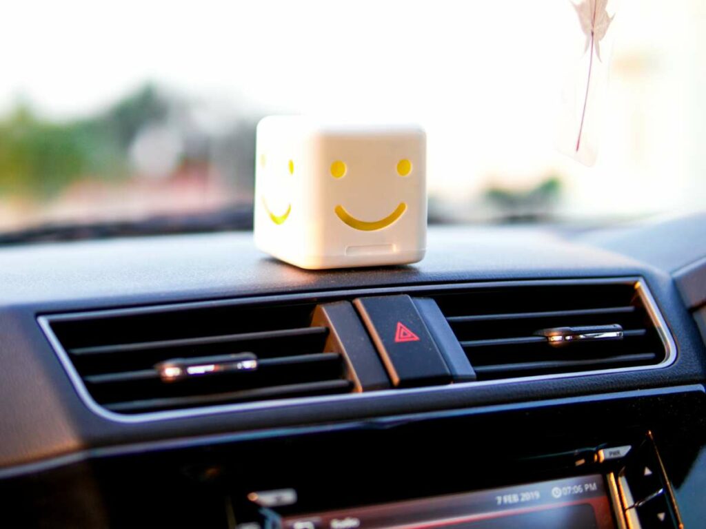 A small, cube-shaped toy with a smiling face sits on a car dashboard.