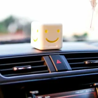 A small, cube-shaped toy with a smiling face sits on a car dashboard.