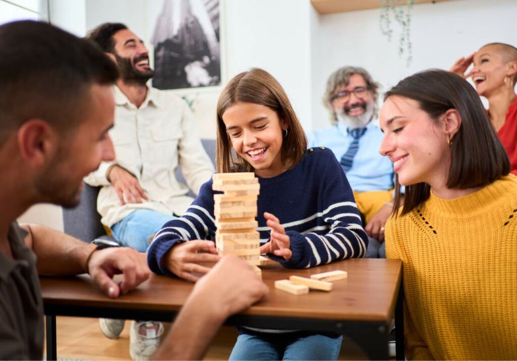 A group of people sits around a table, smiling and laughing while playing a game of Jenga. The focus is on two girls who are placing blocks on the tower.