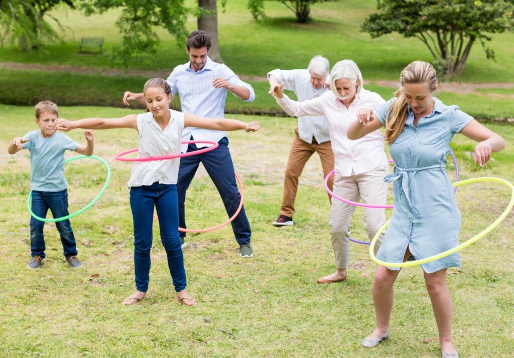 A group of adults and children hula hooping together on a grassy field in a park on a sunny day.