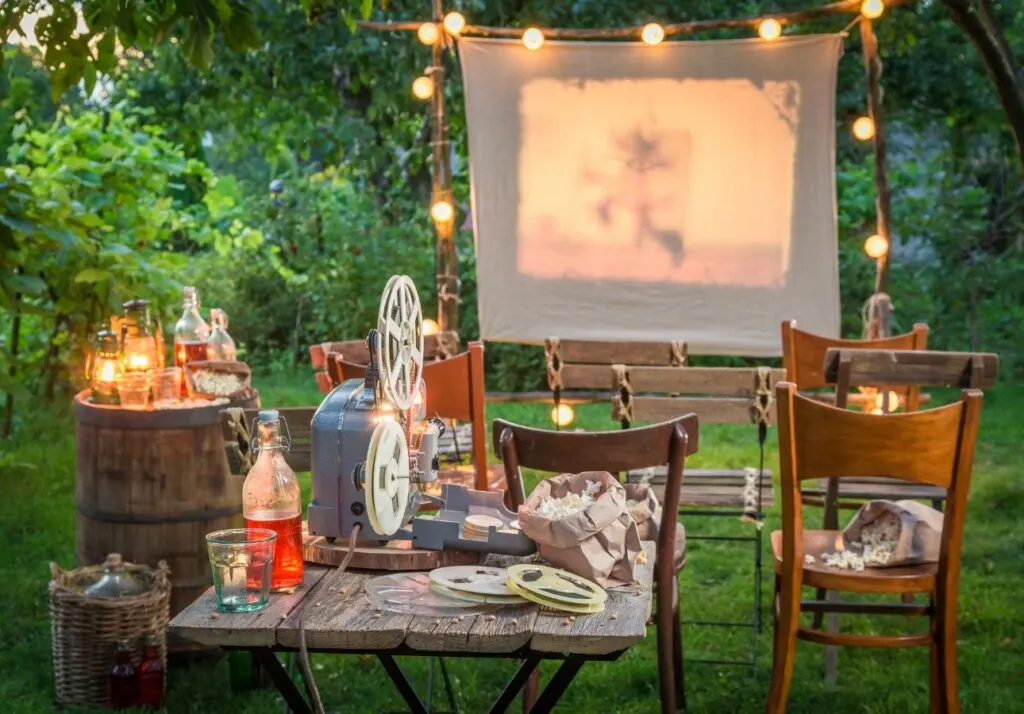 Outdoor movie night setup with a projector, screen, string lights, and a table with snacks in a garden at dusk.