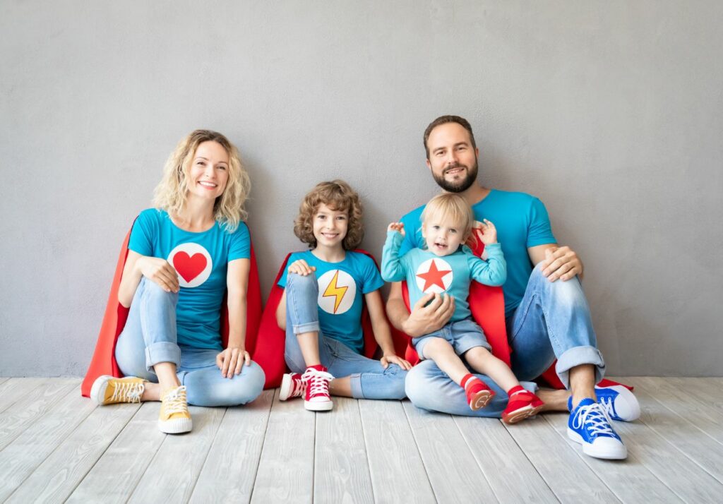 A family of four dressed in superhero t-shirts and capes, sitting and smiling against a gray background.
