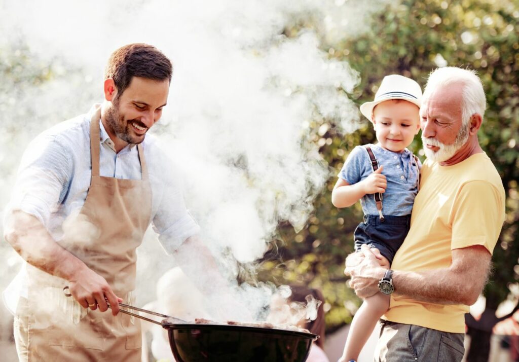 A young man grilling outdoors, holding a toddler, who is watching intently, while an older man stands beside him, smiling contentedly.