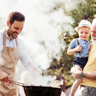 A young man grilling outdoors, while an older man stands beside him holding a toddler.